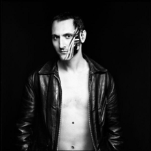 Black-and-white image of Mirwais Ahmadzaï wearing a leather jacket only with its zip open. The left side of his face is painted.
