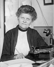 Hopley at her desk, a pencil hand. On her desk in front of her are a typewriter, telephone, and stacks of envelopes.