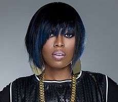 Image of rapper Missy Elliott looking to the camera with her mouth open. She is sporting short black and blue hair, heavy make-up, black shirt with white stripes, and gold earrings and necklace.