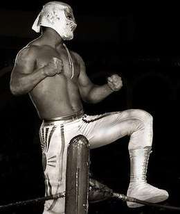 A masked wrestler, Místico, wearing a silver colored mask trunks, posing on the turnbuckles during a wrestling event