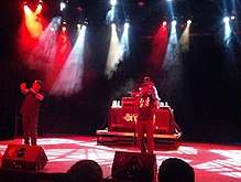 Two African-American men rap into microphones on-stage, as a disc jockey plays music in the background.