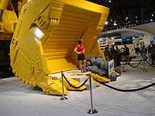 a female model, in a red top and black skirt, standing (with her hands on her hips) in a (bright yellow) bucket of an hydraulic shovel in the exhibit hall of MINExpo 2008. The model is standing behind a velvet rope discouraging trade show attendees from standing in the bucket. Another exhibit is visible in the background as is a group of approximately 12 attendees all but one of which appear to me men.