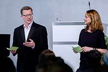 A man and a woman moderate a talk on a stage together.