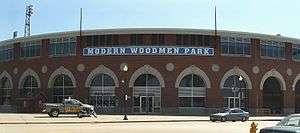 A large brick stadium with many round windows on the bottom and many rectangle windows in groups of four line the top of the stadium. The words Modern Woodmen Park are displayed above the door