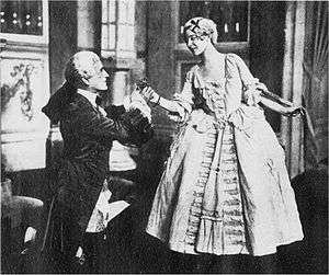Photo of a scene in upper-class 18th century costume, a young man kneeling and grasping the hand of a young woman, who is standing and leaning towards him