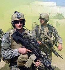 Michael Monsoor, a U.S. Navy SEAL, with a fellow SEAL teammate, dressed in green camouflage uniform loaded with green combat uniforms. Both are carrying firearms and wearing sunglasses. There is a white-colored building and green smoke billowing in the background