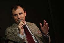 Photo of Jesse Thorn speaking into a microphone. He is caucasian and wearing a coat and tie.