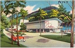 A color image postcard with a park lodge in the background and a red convertible in the foreground.