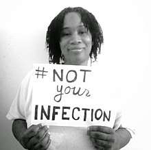 Half-length portrait of a Black woman in a white shirt holding a sign with the hashtag #NotYourInfection