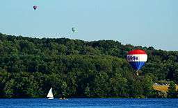 Three hot air balloons over a lake with a sailboat and forested shore