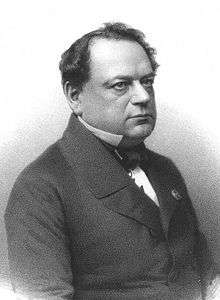 Photograph of a lithograph. It shows the torso and head of a middle-aged man who is apparently seated and is looking off to the reader's right. He is dressed formally, and is wearing a coat, a white shirt, and a tie. There is a metal pin on his lapel. He has black hair, is somewhat balding, and is slightly overweight.