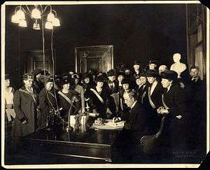 A man sits at a desk signing a document while a large group of women watches