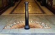 Close-up photograph of a black Victorian-style bollard in the centre of the arcade walkway. A semi-circular mosaic on the floor spells out the letters "Queen's Arcade" around the bollard.