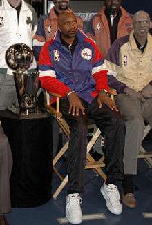 A black man wearing a blue and red jacket sits on a wooden chair. Several others and a trophy sit next to him