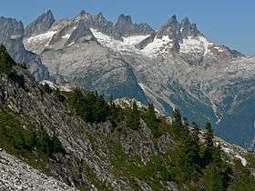 A range of sharp mountain peaks. The lower reaches have snow on them. In the foreground, a wooded slope descends from left to right.