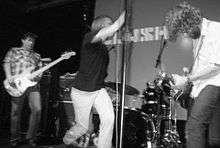 A photo of a rock band, Mudhoney, at a live show. The photo is blurred from the onstage motion. From left to right are the electric bassist, singer and guitarist.