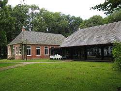 A red brick building and a glass building, both having grey tile roofs, with green grass in the front and green trees in the background