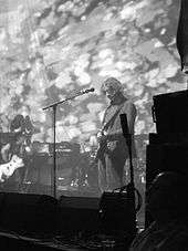 A monochrome image of three musicians performing onstage against a psychedelic backdrop.
