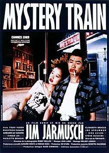A young Japanese couple are sitting on a dimly-lit street outside a restaurant with a neon sign. The woman is looking off camera and resting against the man, who is downcast with a cigarette in his mouth; both are wearing bright red lipstick. Superimposed above the couple is the title MYSTERY TRAIN, with credits in French below the image.