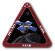 An artwork of a spacecraft hovering above an asteroid, enclosed in an equilateral triangle with thick, red border. The words "JHU/APL", "NASA", and "NEAR" are printed in bold white font, on the left, right, and bottom sides of the triangle's borders.