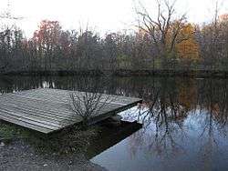 A wooden dock on the shore of a small lake in which autumnal trees are reflected