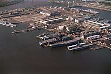 An overhead view of a large shipyard. Various ships can be seen tied up, with several structures visible within the yard.