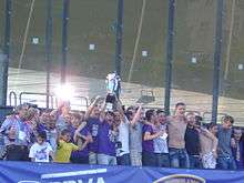 A group of individuals, some with arms round each other's shoulders, standing in a row, with two of them holding a silver trophy in the air (29 May 2011, after the match vs Domžale).