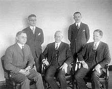 A black-and-white photo of five men wearing business attire. Two are standing and three are seated.