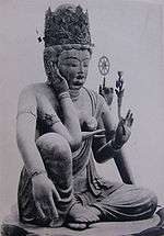 Three-quarter view of a seated statue with six arms. The left leg is bend, touching the ground along its whole length. The right leg is also bend, but with the knee up. The right foot is above the left foot. One of the right hands is touching the right ear, another is lifting a jewel-shaped object in front of the breast. One of the left hands carries a flower, another a small wheel-shaped object.