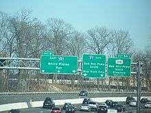 A multilane freeway with a display of three green signs over the road. The left one reads exit 8 east Interstate 287 White Plains Rye three downward arrows, the middle one reads Interstate 87 south Saw Mill Parkway south New York City two downward arrows, and the right one reads exit 8A New York State Route 119 Saw Mill Parkway north Elmsford right lane.