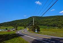 A two-lane paved road winding through countryside from just right of the camera, down the center of the frame, towards a hill covered with green trees under a blue sky with some small clouds in it. On the far side of the road there is a sign with the number 22 on it; below it is a white on blue sign with "Be Prepared to Stop" on it in capital letters. Telephone wires enter the image from top left, connecting to a wooden pole at the center