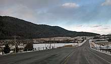 A two-lane paved road curves rightward away from the viewer toward a rise in the center of the image amid a snowy landscape of fields and woodlots under mostly cloudy skies, with a ridge of hills on the left