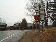 A pair of highways intersect in front of a pair of cabins in a rural area. The highway in the foreground ends here, forcing drivers to turn left or right. A nearby sign assembly indicates that NY 28 west is to the left while NY&nbsp;28N east and NY&nbsp;30 north are to the right.