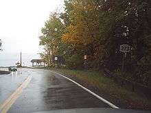 A two-lane highway turns to the left as it approaches a body of water. To the right of the highway is a NY 373 shield.