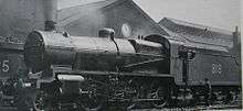 Side-and-front view of a modified version of the N&nbsp;class on shed. The distinguishing feature from normal N&nbsp;class locomotives is the tall cylindrical chimney on the smokebox. A member of the crew is standing next to the tender.