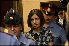 A serious-looking woman with a brown bob haircut and plaid shirt, is led into a courtroom between two uniformed officers.