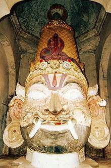 A big wooden male head with a moustache, bushy brows, Vaishnava tilak, fangs protruding from the end of the mouth, big eyes and ears and wearing a conical crown with a cobra on its front and  another five-headed cobra is emerging from behind
