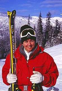 A smiling female skier wears a red winter jacket, white gloves, and white goggles over of a black winter cap. She holds a pair of skis in her right hand and a pair of ski sticks in her left hand. Behind her, a snowy pine-covered landscape.