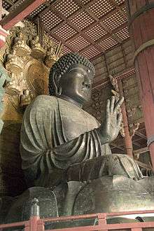Three-quarter view and worm's-eye view of a seated statue. The palm of his right hand faces forward. The folds in his clothing are deeply sculpted. Behind the head of the sculpture there is a halo decorated with sitting statues. Color picture.