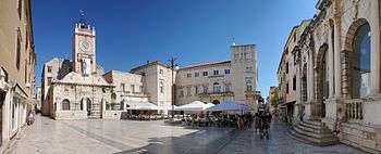 A square in a town which is surrounded by medieval buildings with towers