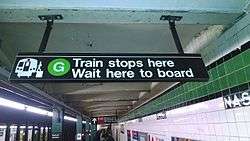 An overhead sign at an underground "G" train station. The sign's text says, "'G' train stops here. Wait here to board". There is an icon of a train on the left side of the sign, next to the text.