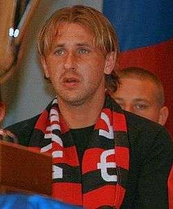 A white blond male seating on a stand with a black and red FC Khimki scarf around his neck.
