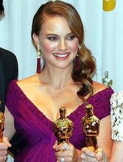 An image of a smiling woman with light brown hair in her 20s. She is wearing a purple dress and is holding a golden statue.