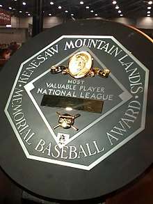 A black circle with an octagonal silver plaque in the middle. The edge of the plaque reads "KENESAW MOUNTAIN LANDIS MEMORIAL BASEBALL AWARD". In the middle of the octagon is a baseball diamond which contains, from the top, Judge Landis' face in gold, "Most Valuable Player", the winner's league, his name in a gold rectangle, and his team.
