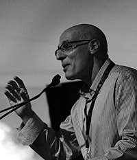 Black and white photograph of Gordon Korman, author of young adult fiction, speaking at the National Book Festival in September 2011. Photograph depicts Korman in profile view, facing left and speaking into a microphone. His right hand is raised to approximately shoulder height, palm facing the audience, with fingers slightly closed as if grasping an invisible ball.