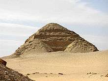 A photograph of the pyramid. The exterior consists of heaped layers of rubble draped over the stepped pyramid core. The stepped blocks protrude out from underneath.