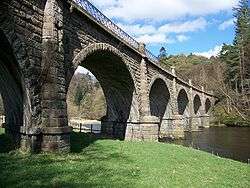 A handsome bridge, built from multiple semi-circular arches of buff sandstone spans the river in picturesque surroundings. Four of the piers are in the water and the helical courses of stone attest to its oblique construction. Atop the parapet is a cast iron railing.