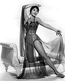 Dancer posing expressively in an ornate leotard and sheer fabric costume, one knee resting on an upholstered bench