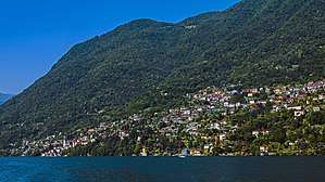 A large settlement consisting mostly of light-colored structures with orange roofs rising up the lower slopes of forested mountains seen from a body of water under a blue sky