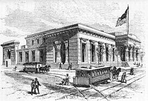 An Egyptian-Revival-style buidling on a street corner, with doric columns on its façade and a large flag flying atop a mast. Pedestrians are seen navigating two horse-drawn trams, which travel on lines that bisect intersecting streets.